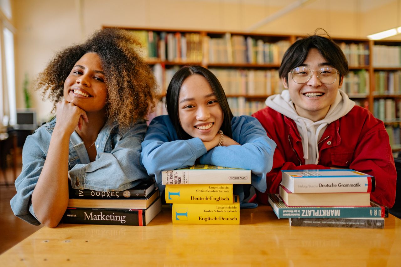 About Three happy students in a library with language and marketing books.